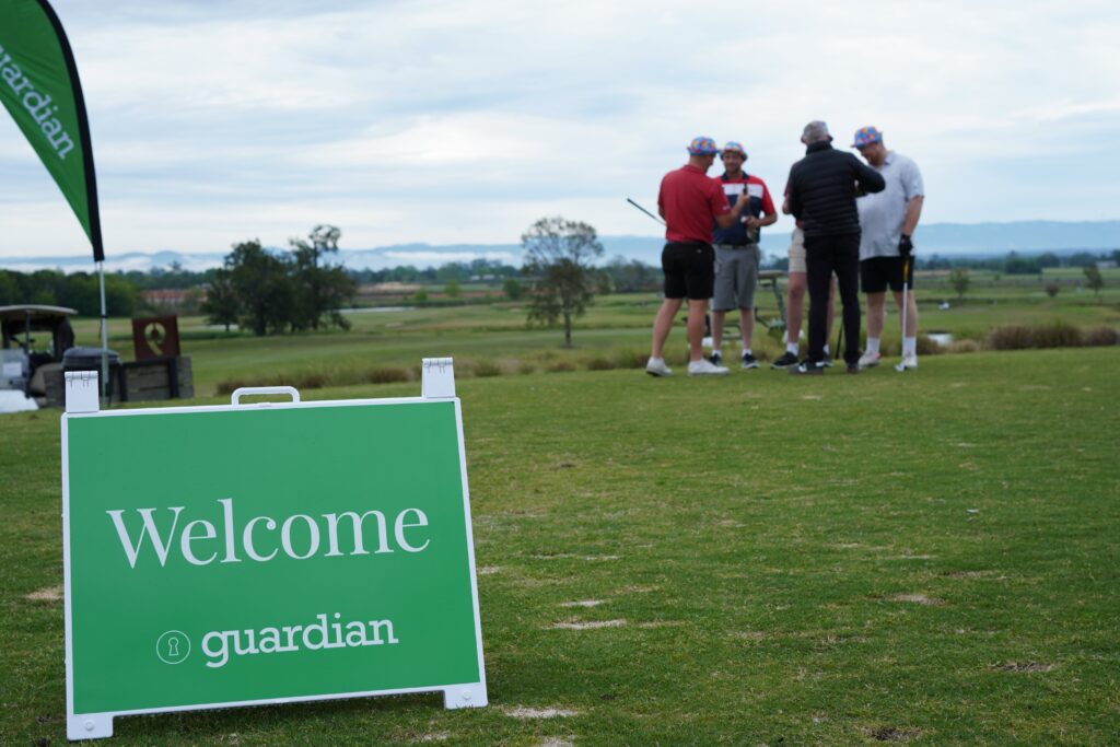 A group of men are talking before starting a round of golf. A Welcome sign is in the foreground.